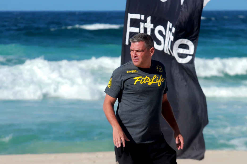 Fabiano José Moraes com camiseta FITSLIFE e bandeira na praia.